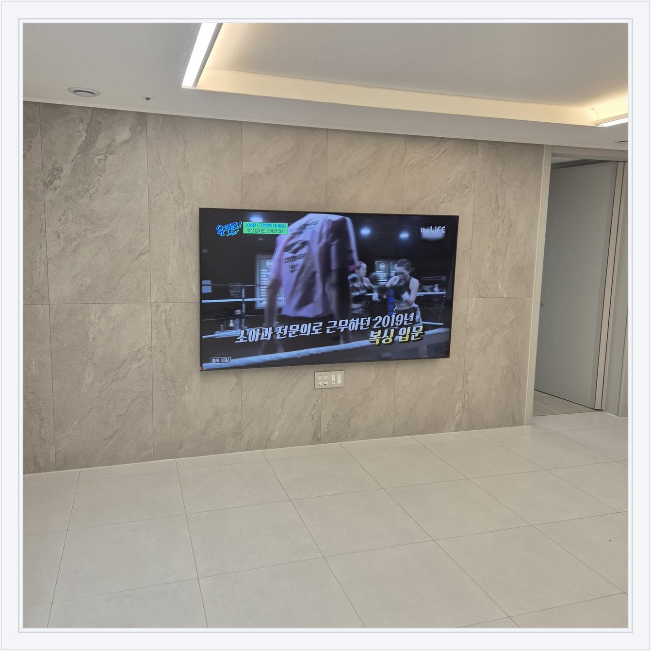 석재 느낌 벽면을 정면에서 본 TV, 넓은 바닥 여유와 조명 라인이 동시에 보입니다