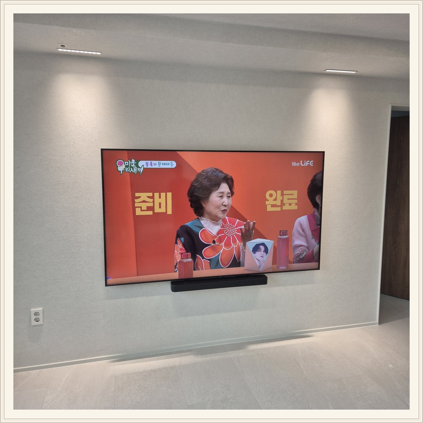 조명이 비추는 미색 벽과 사운드바, 화면 아래 가로로 놓인 검은 음향기기가 선명합니다