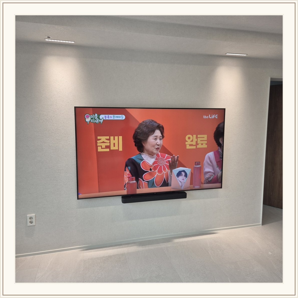 조명이 비추는 미색 벽과 사운드바, 화면 아래 가로로 놓인 검은 음향기기가 선명합니다