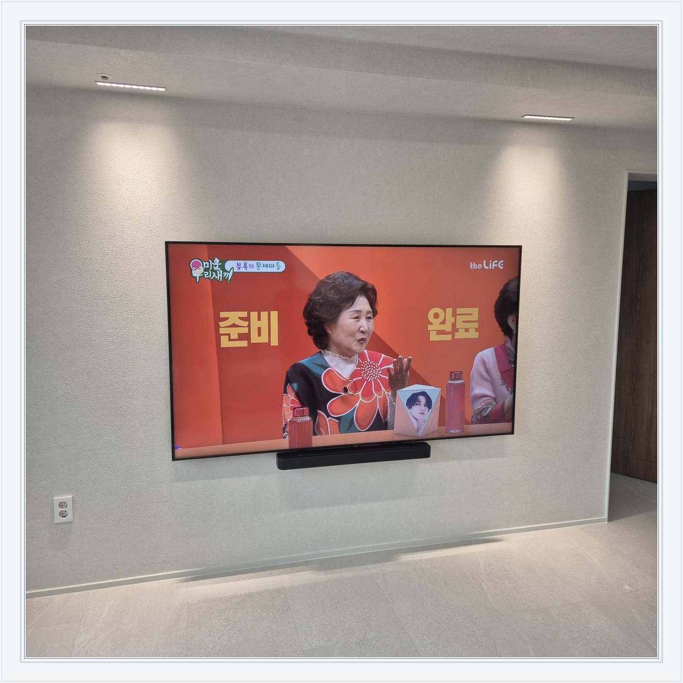 조명이 비추는 미색 벽과 사운드바, 화면 아래 가로로 놓인 검은 음향기기가 선명합니다