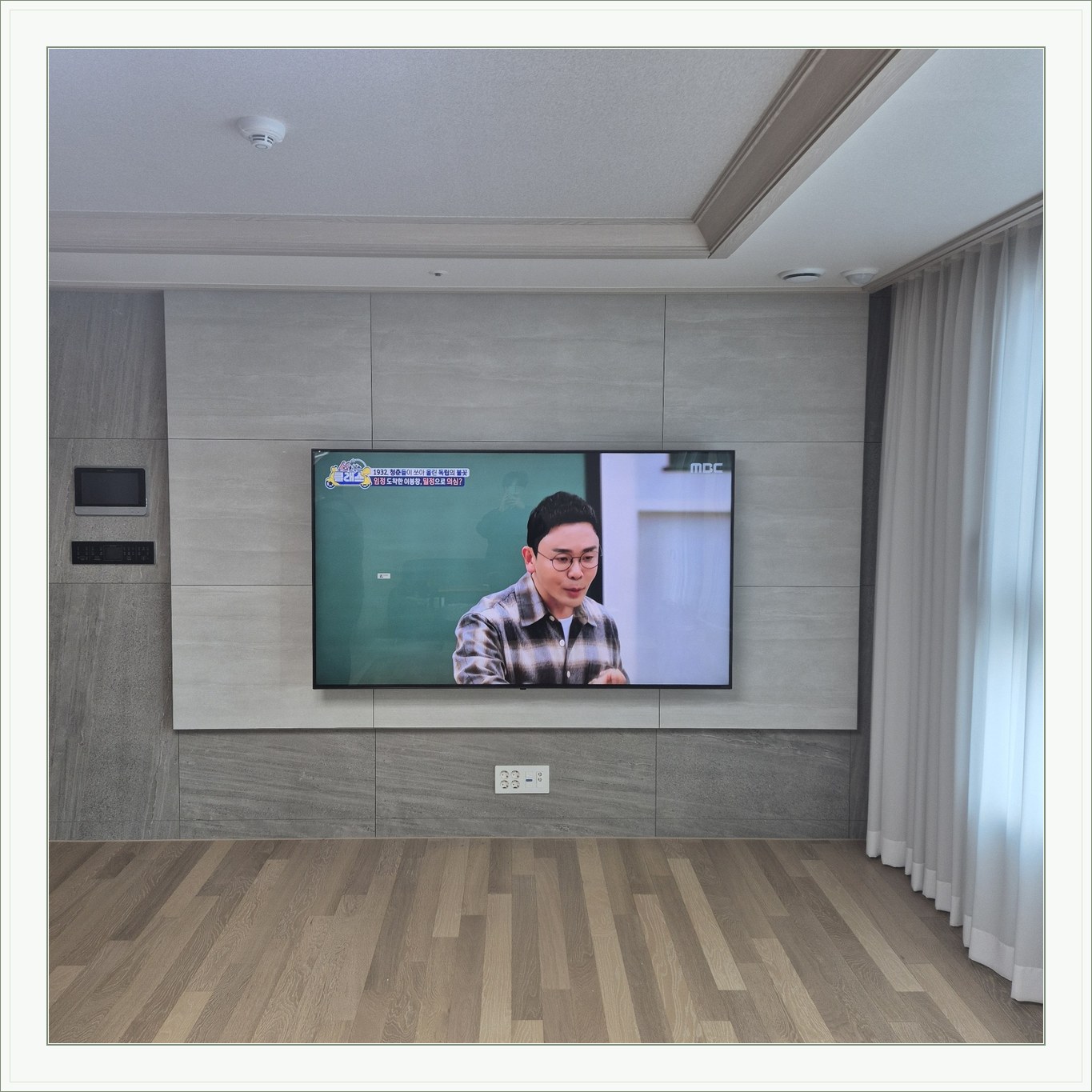거실 회색 패널과 벽걸이 TV, 오른쪽 커튼 옆으로 화면이 중심에 놓여 있습니다